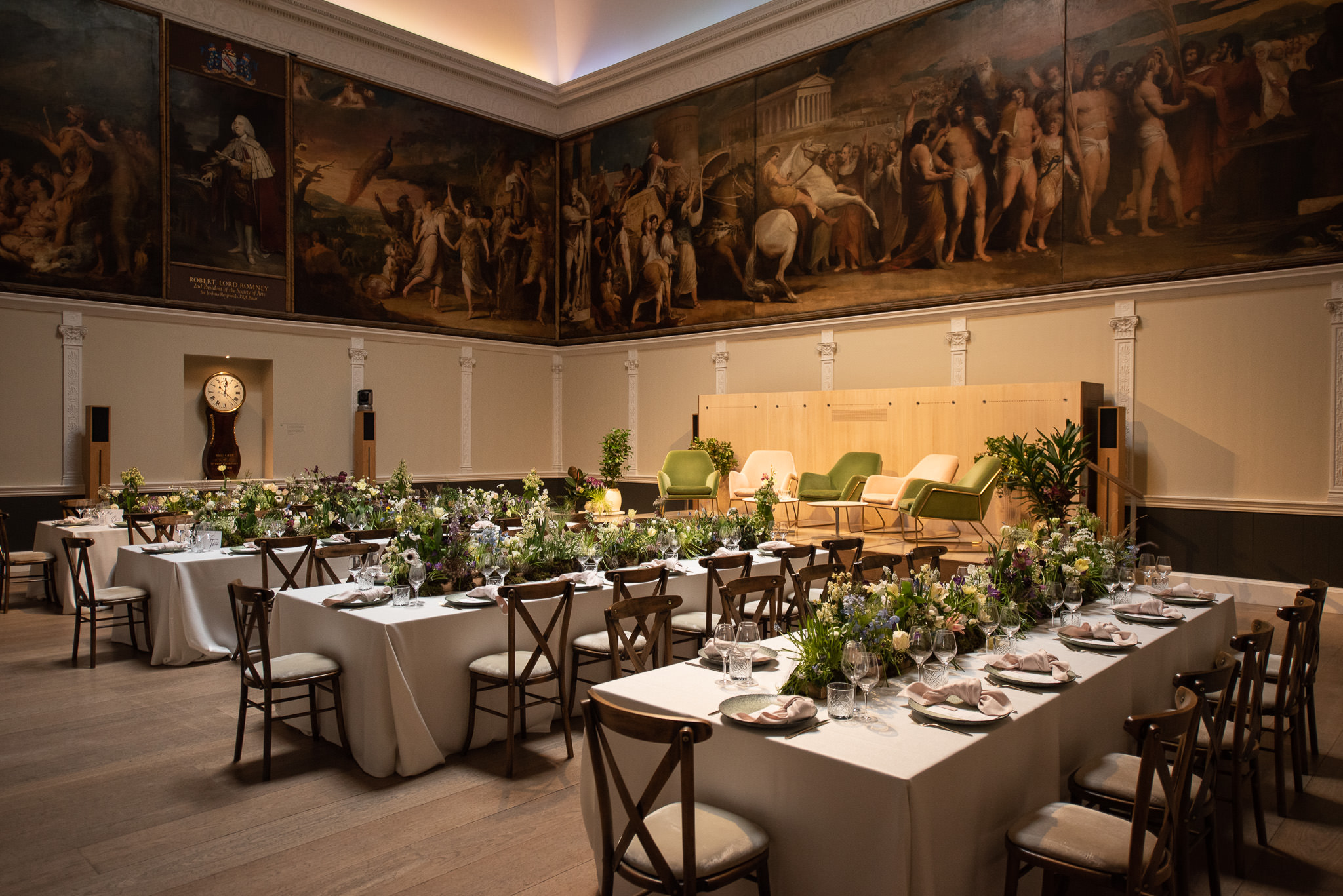 Elegant dining hall with three long tables, adorned with floral centerpieces and place settings. Large historical paintings adorn the walls. Soft, warm lighting illuminates the room, creating a sophisticated ambiance.