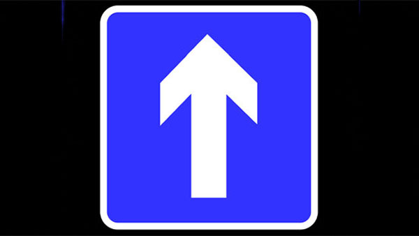 Blue road sign with a thick white arrow pointing upward, indicating one-way or straight ahead. The sign has a white border and is set against a black background.