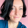 A person with shoulder-length dark hair smiles at the camera. They are wearing a blue top and are in front of a pink background.