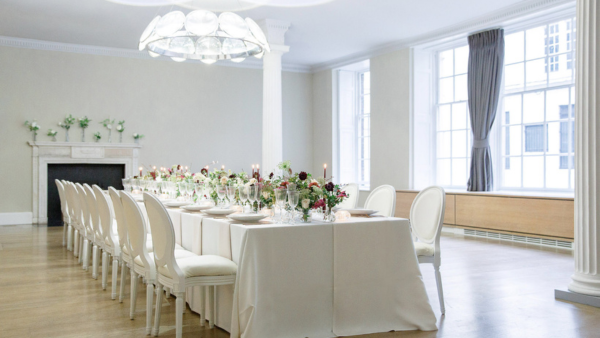 A bright dining room with a long, elegantly set table featuring white chairs, floral centerpieces, and lit candles. Large windows with gray curtains line the walls, and a white fireplace with floral decor is on the left.