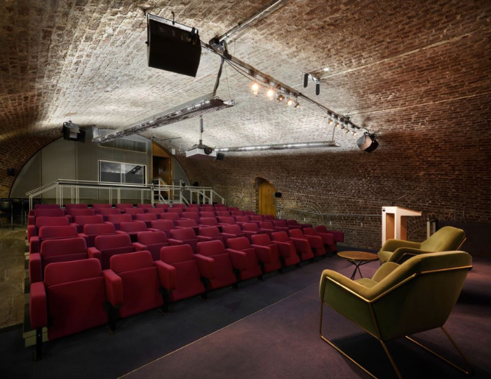 An auditorium with red cushioned seats facing a stage under a vaulted brick ceiling. Two green chairs and a podium are on the stage, lit by overhead lights. A large screen is in the background.