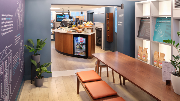 Modern coffee shop interior with blue walls and light wood flooring. A long wooden table with orange cushioned benches is in the foreground. The counter area features pastries, snacks, and coffee machines. Plants and shelving add to the decor.