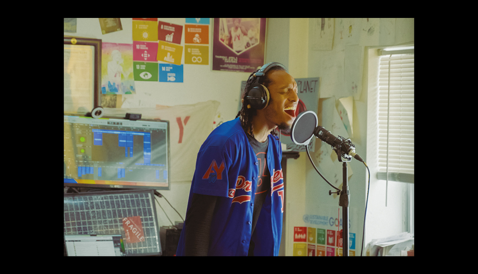 Person wearing headphones and a blue sports jersey sings into a microphone in a music studio. The room has posters on the wall, a computer with audio software open, and a window with blinds partially drawn.