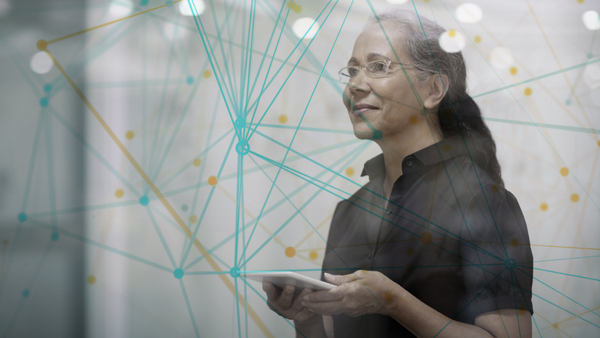 A person holding a tablet stands behind a transparent screen with interconnected lines and dots, symbolizing a network. The person is wearing glasses and a dark shirt, looking thoughtful. The background is blurred.