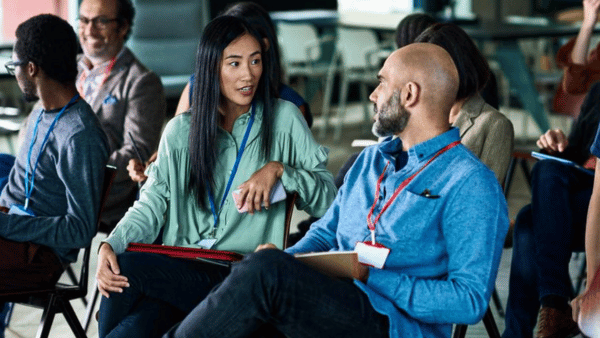 A diverse group of people sitting in chairs, engaged in conversation. Two individuals in the foreground, wearing lanyards, are actively discussing. Others in the background appear to be listening or participating in similar discussions.