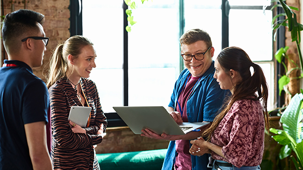 A group of four people in casual attire are gathered in an office setting with large windows and plants. They are smiling and engaged in conversation. One person is holding a laptop while another holds a tablet.