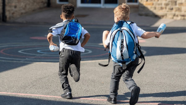 Two young boys with backpacks are running on a school playground. One carries a water bottle. The sun casts shadows on the pavement, and colorful chalk drawings are visible in the background.