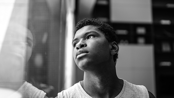 Black and white image of a person looking thoughtfully through a window. Their reflection is visible on the glass. The background is softly blurred, focusing attention on the contemplative expression.