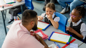 A teacher crouches at a desk, engaging with two young students. The children sit with whiteboards, one smiling and looking at the teacher, the other wearing glasses. The classroom has blue chairs and various school supplies scattered on the desk.
