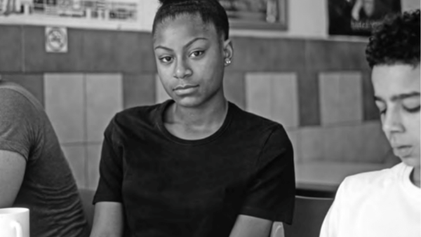 Three people are seated in a cafe. A person in the center is wearing a black t-shirt and has their hair up, looking directly at the camera. The person on the right is wearing a white shirt and looking down. The image is in black and white.