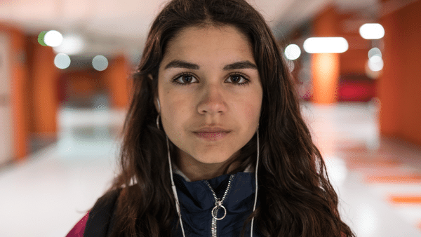 A young woman with long brown hair looks directly at the camera. She is wearing a dark blue jacket and has white earphones. The background features a brightly lit, modern hallway with orange accents.