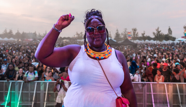 A woman stands confidently in front of a large crowd at an outdoor event. She is wearing sunglasses, a white tank top, and colorful beaded necklaces. Her right fist is raised in the air, and the audience is visible in the background.