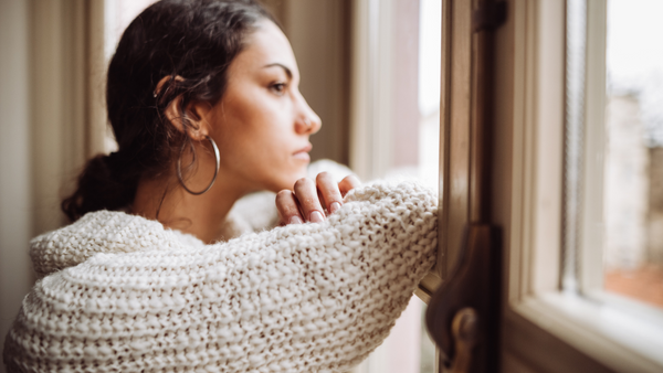 A woman with long hair and large hoop earrings gazes thoughtfully out of a window. She is wearing a chunky knit sweater and leaning on her hand, with an expression of contemplation. The scene is cozy and introspective.