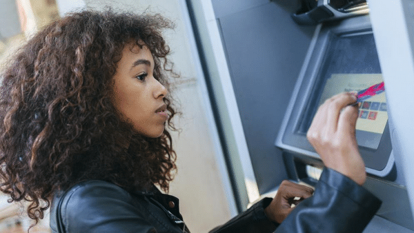 A woman with curly hair wearing a black leather jacket uses an ATM. She is inserting a card into the machine while looking at the screen.