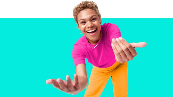 A smiling person with short curly hair reaches out with open hands. They are wearing a bright pink shirt and yellow pants, set against a turquoise and white background.