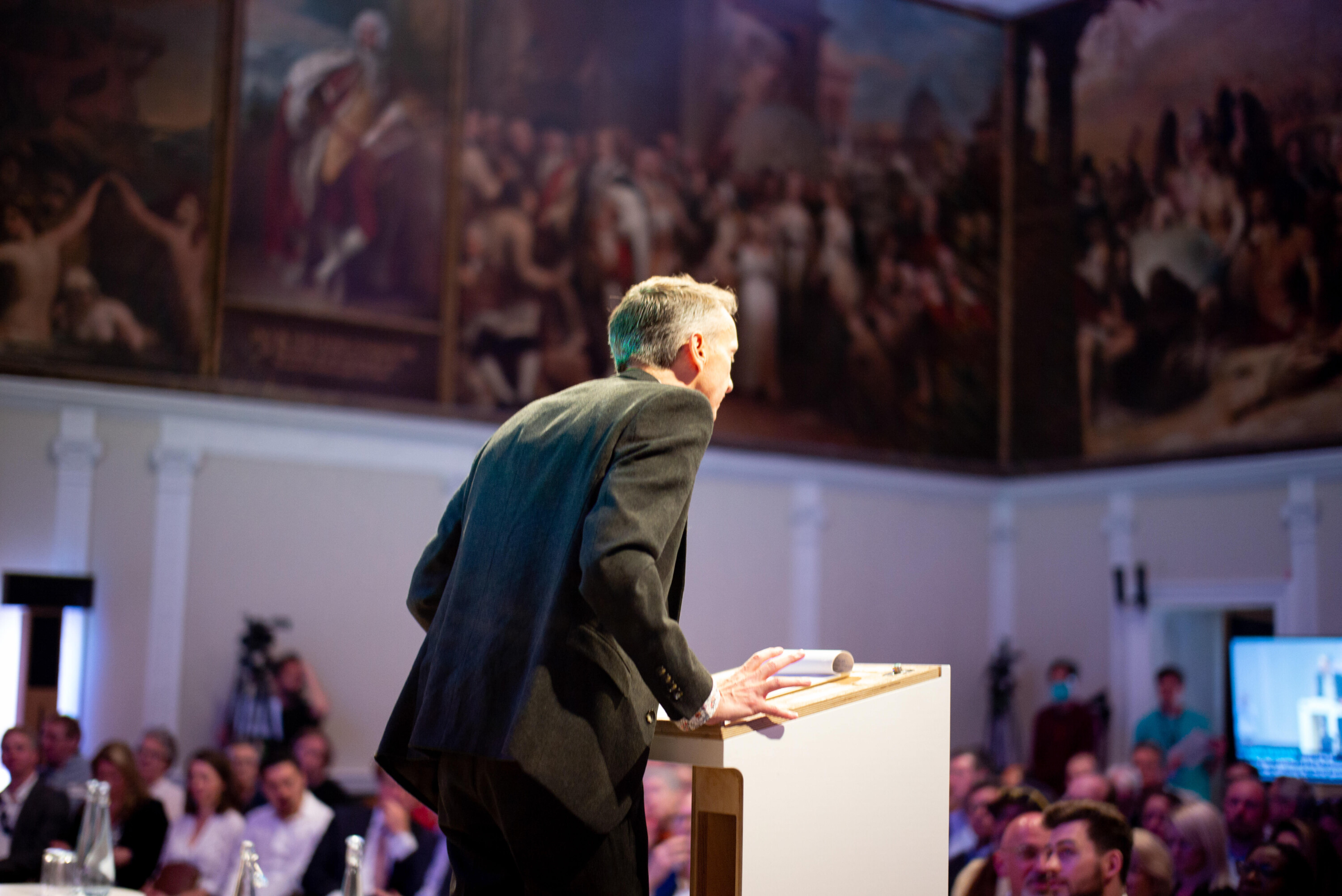 A speaker stands at a podium addressing a large audience in an ornate hall with classical paintings on the walls. The room is filled with seated attendees, and there is a microphone on the podium.