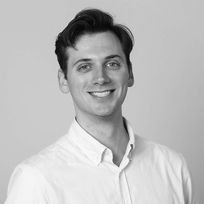 A man in a white button-up shirt smiles at the camera. The photo is in black and white, with a plain background.
