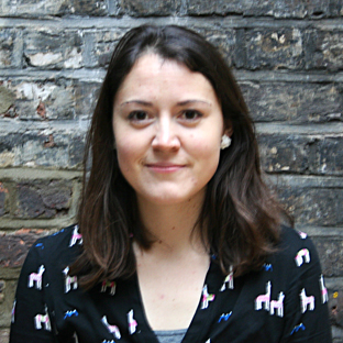 A person with shoulder-length brown hair is standing against a brick wall. They are wearing a black sweater with a colorful pattern.