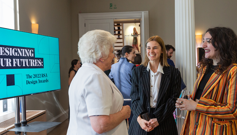 Three women talking and smiling at an indoor event beside a screen displaying "Designing Our Futures: The 2022 RSA Design Awards." A woman in a striped jacket holds a drink. Other people are visible in the background.