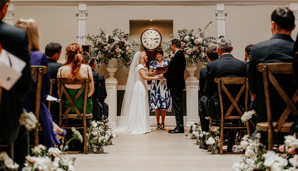 A bride and groom stand holding hands during a wedding ceremony. A person stands between them officiating. They are surrounded by seated guests and floral arrangements, with a large clock in the background.