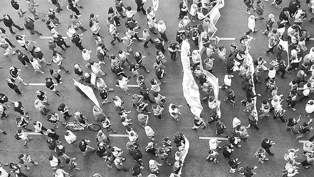 Aerial view of a large crowd gathered for a protest. People are marching on a street, holding signs and banners. The image is in black and white, highlighting the density and movement of the participants.