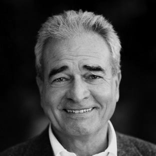 A grayscale portrait of an older man with short, gray hair. He is smiling, wearing a white shirt and a dark blazer. The background is blurred, keeping the focus on his face.