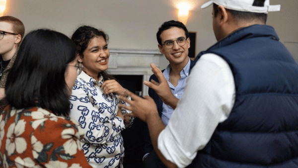 A group of people are engaged in a lively conversation indoors. One person in a patterned shirt is smiling, while another in a blue shirt and glasses looks on attentively. A person in a cap and vest is gesturing animatedly.