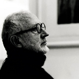 Black and white profile photo of an older man with glasses and a beard. He is wearing a dark turtleneck sweater and looking off to the side, with a framed picture blurred in the background.