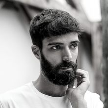 A black-and-white photo of a person with short, dark hair and a beard, wearing a light-colored shirt. They are looking to the side with one hand touching their chin. The background is slightly blurred, suggesting an outdoor setting.