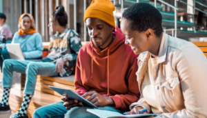Two people sit on a bench; one wearing an orange beanie and hoodie is using a tablet, while the other with short hair holds a notebook and pen. In the background, three people are sitting, one with a laptop, in a relaxed setting.