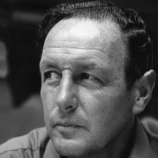 Black and white portrait of a person with short, dark hair, looking to the side with a contemplative expression. The background is blurred.