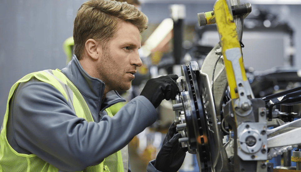 A person wearing a yellow safety vest and black gloves is focused on adjusting machinery. They are working in an industrial setting, surrounded by equipment. The person appears attentive, concentrating intently on the task.
