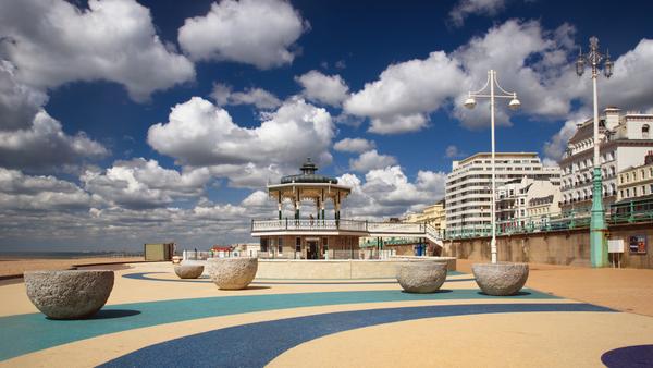 A seaside promenade features a decorative bandstand, large stone planters, and colorful paving patterns. Tall buildings line one side, with a clear blue sky and scattered white clouds above.