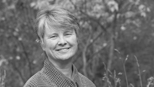 A grayscale portrait of a smiling person with short hair, wearing a textured jacket. They stand outdoors in front of blurred foliage and branches.