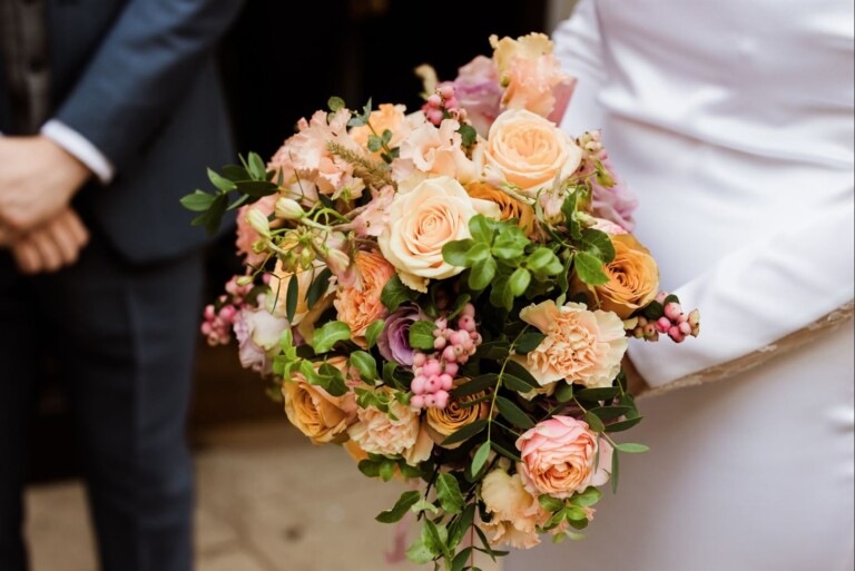 A bride holding a bouquet of pastel roses, pink berries, and green foliage. The bouquet includes peach, pink, and cream roses with a touch of purple. A person in a suit stands beside the bride.