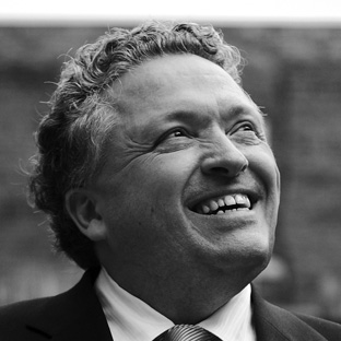 Grayscale image of a person with curly hair, wearing a suit and tie, smiling while looking upwards.