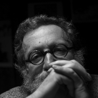 Black and white close-up of a person with glasses and a beard, hands clasped under their chin. The lighting is dim, focusing on the contemplative expression.