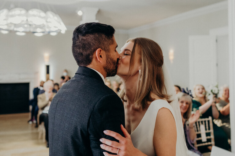 A couple shares a kiss at their wedding ceremony. The groom wears a dark suit, and the bride is in a white dress. They are surrounded by guests seated on chairs, some smiling and applauding. The room is elegantly decorated with soft lighting.