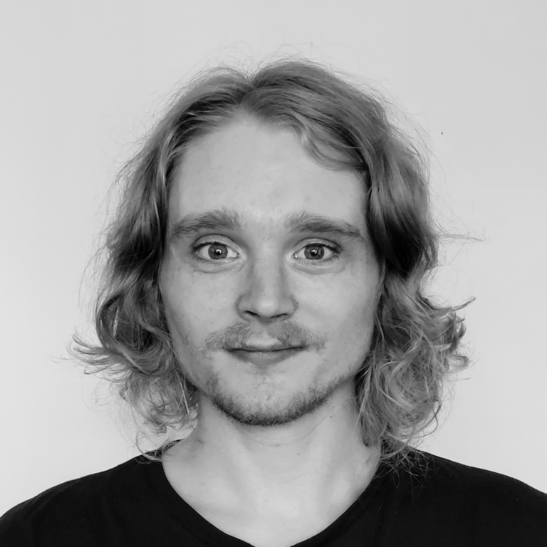 A black and white portrait of a person with wavy shoulder-length hair and facial hair, wearing a simple dark shirt. They face the camera against a plain background, creating a minimalist and focused composition.