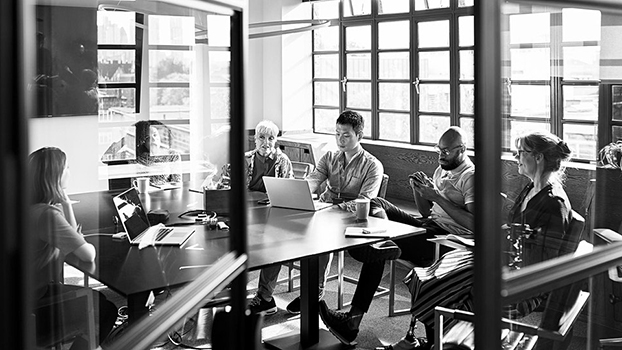 A black and white photo of a diverse group of five people sitting around a conference table in an office setting. They are engaged in conversation, with some using laptops and others looking at a smartphone. Large windows in the background let in natural light.