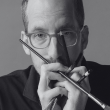A person with glasses holds several writing instruments, including pens and pencils, close to their face. They wear a dark shirt and have a contemplative expression. The image is in black and white, focusing closely on their upper body and hands.