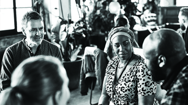A group of diverse people sitting in a circle and having a conversation in a cozy, light-filled room. The atmosphere appears warm and engaging, with several plants and people in the background.
