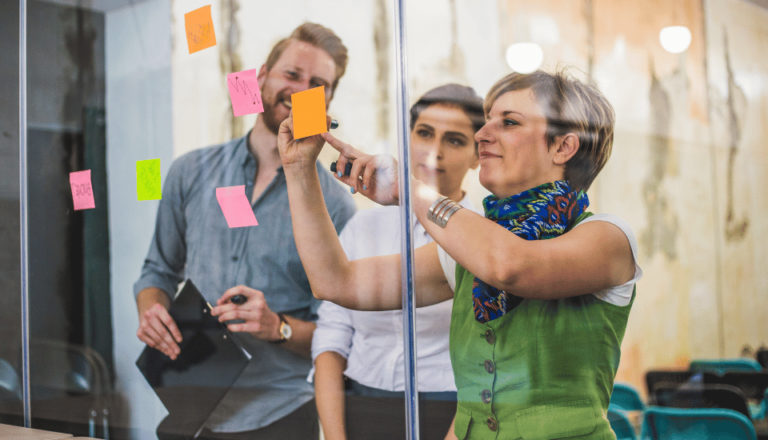 A group of three people collaborating in an office setting. A woman in the foreground is writing on colorful sticky notes placed on a glass wall, while two others stand behind her watching attentively. They appear engaged and focused.