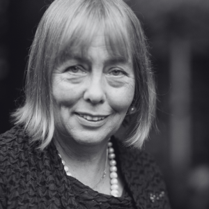 Black and white portrait of a smiling older woman with shoulder-length hair. She wears a textured jacket and a pearl necklace. The background is softly blurred.