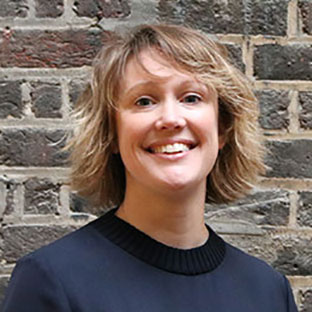 Smiling person with short blonde hair wearing a dark top stands in front of a brick wall.