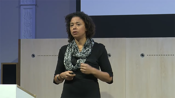 A person with curly hair is speaking on stage, holding an object in their right hand. They are wearing a black top and a patterned scarf. The background features a light-colored wall and part of a projector screen.