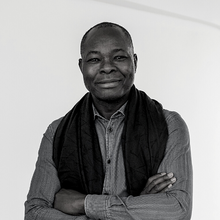 A person stands with arms crossed, wearing a dark scarf and a button-up shirt. The background is a plain white wall.