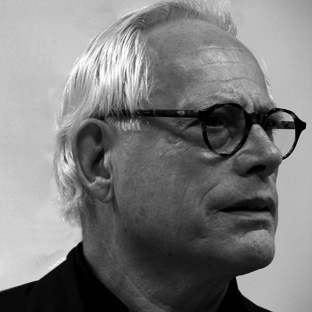 A black and white image of an older person with short, light-colored hair and glasses. They are wearing a dark top and looking to the side with a thoughtful expression. The background is plain.