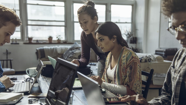 Four people are gathered around a table working on laptops and tablets in a well-lit room. One person is standing, pointing at a screen while the others sit and focus on their devices, suggesting a collaborative work environment.