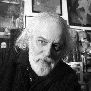 Dionysis Fotopoulos with long white hair and a beard gazes intently at the camera. He is sitting in a room filled with various framed artworks and objects in the background, creating an eclectic and artistic atmosphere. The image is in black and white.